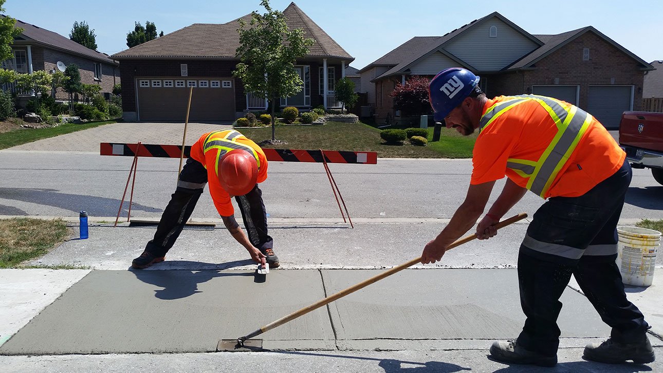 2 men smoothing out concrete for sidewalk next to a street in front of a residential home